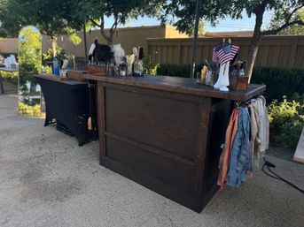 Rustic wooden outdoor bar and prop station with jars of feathers, colorful fabric ribbons hanging at the side, small American flags in a white vase, a tall mirror and trees along a backyard fence for an outdoor event.