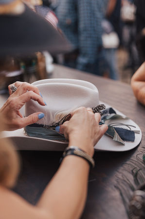 Hands with blue nail polish attaching feathers and fabric ribbons to a light gray felt hat on a wooden table, styling a custom hatband at a craft workshop.