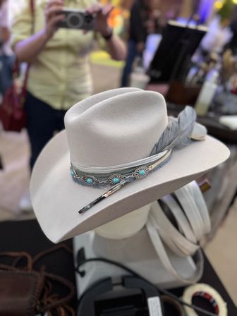 Light gray felt Western hat with turquoise-stone concho band, feather and decorative hatpin on a mannequin, displayed at an artisan market booth