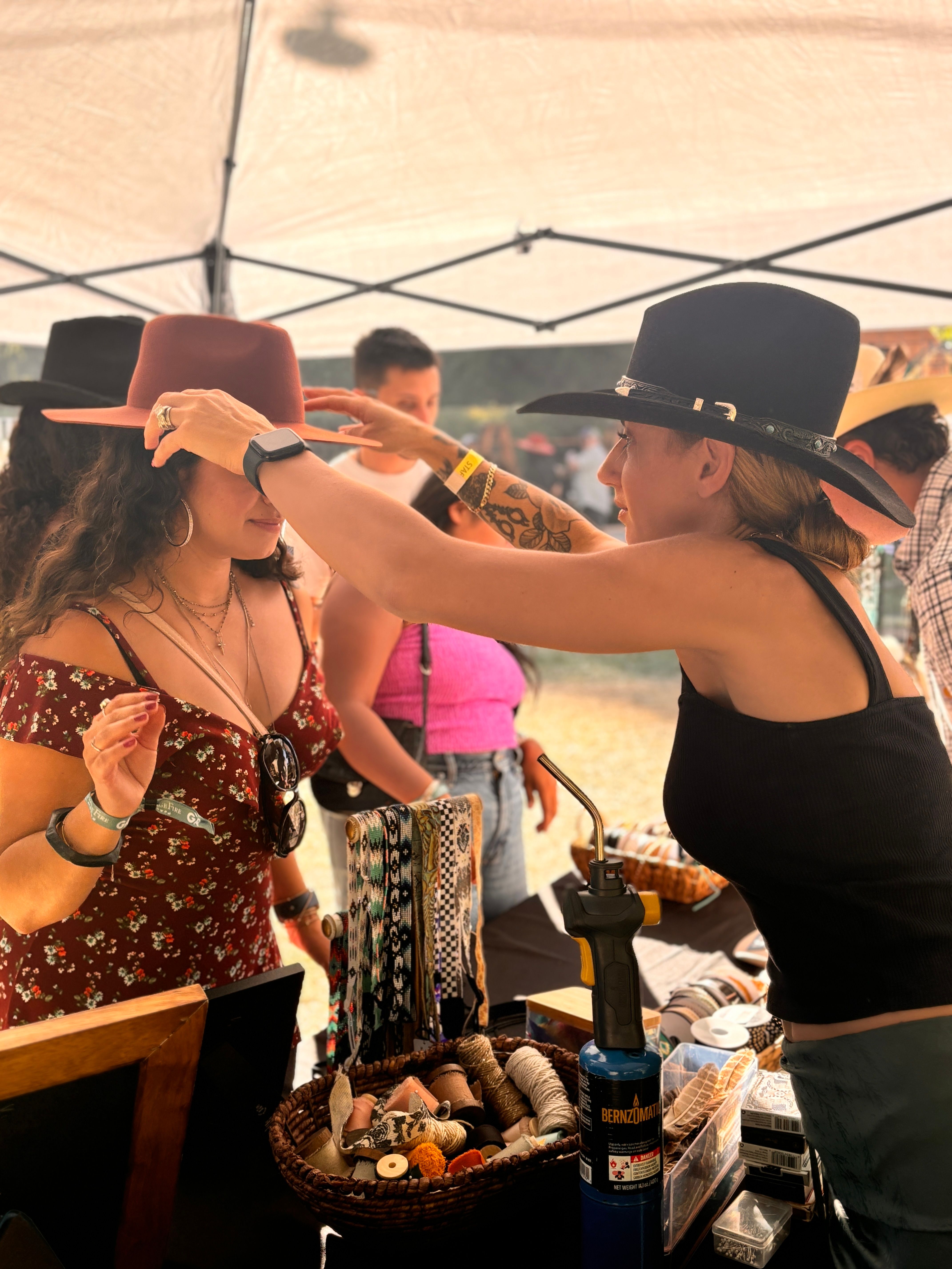 Vendor adjusts a wide-brim hat on a shopper at an outdoor artisan market booth under a canopy, table of woven hatbands, leather trims and craft tools in the foreground.