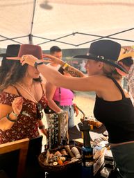 Vendor adjusts a wide-brim hat on a shopper at an outdoor artisan market booth under a canopy, table of woven hatbands, leather trims and craft tools in the foreground.