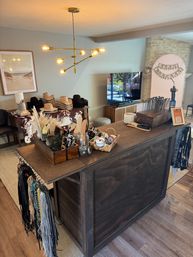 Cozy indoor boutique display with a wooden checkout counter, boho accessories and tools, stacked western hats on cowhide, modern brass chandelier overhead, and a birthday banner in the background