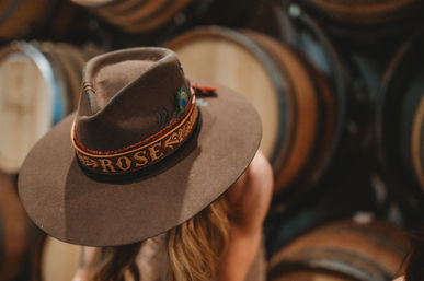 Brown wide-brim felt hat with decorative band and peacock feather worn by a person in a rustic wine cellar with stacked oak barrels in soft focus.