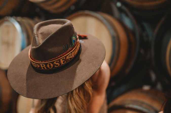 Brown wide-brim felt hat with decorative band and peacock feather worn by a person in a rustic wine cellar with stacked oak barrels in soft focus.
