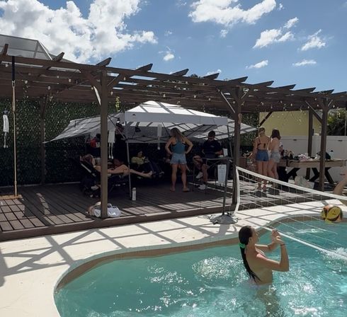 Sunny backyard pool party — people playing water volleyball in a small turquoise pool beside a wooden pergola with a canopy, loungers, string lights and a deck picnic area.