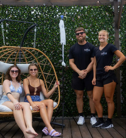 Two smiling friends in swimwear and sunglasses lounging in a rattan hanging egg chair on a wooden patio with a green ivy wall, flanked by two wellness staff in black shirts and an IV hydration pole.