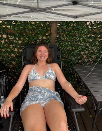 Smiling woman relaxing on a black reclining lounge chair under a canopy in a leafy outdoor patio, wearing a blue-and-white patterned bikini and matching sarong.