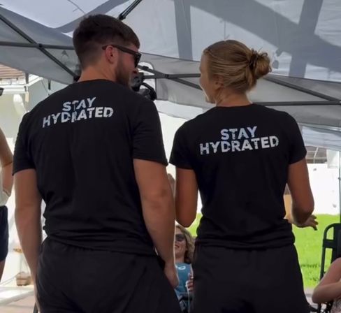 Two people seen from behind under a canopy at an outdoor backyard gathering, both wearing black T‑shirts printed with the slogan "STAY HYDRATED".