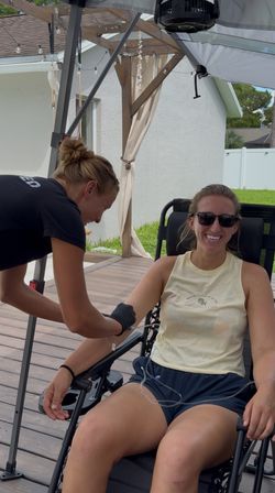 Smiling woman in sunglasses reclines on a backyard wooden deck under a pergola with string lights as a gloved caregiver sets up IV tubing in her arm.
