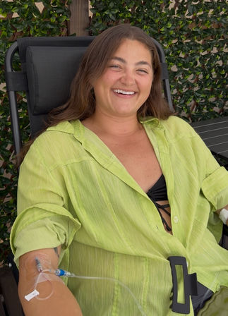 Smiling woman in a lime-green shirt relaxing in an outdoor lounge chair with an IV infusion in her arm and a leafy privacy screen background