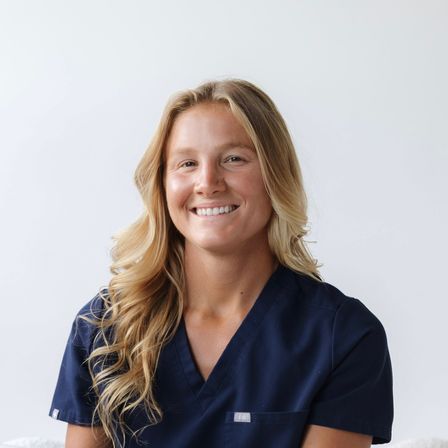 Friendly smiling blonde healthcare professional in navy scrubs — clean studio headshot for medical or clinic profile.