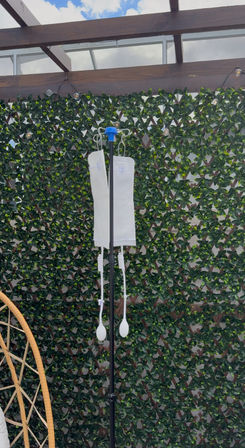 Outdoor patio IV stand: black pole holding two white hydration pouches with squeeze bulbs, set against a green ivy trellis under a wooden pergola and blue sky.