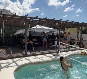 Sunny backyard pool scene with people in swimwear playing pool volleyball, pergola-shaded deck with lounge chairs and a picnic table for a relaxed summer gathering