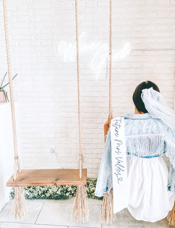 Bride-to-be wearing a veil and fringed denim jacket labeled "Wifey" with a "Future Mrs." sash, seated on a rustic wooden indoor swing against a white brick wall with neon sign glow and low floral greenery.