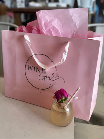 Pink gift bag with crinkled tissue paper and satin handles on a table, paired with a pale cocktail in a stemless glass garnished with a pink rose and striped straw — playful brunch gift scene