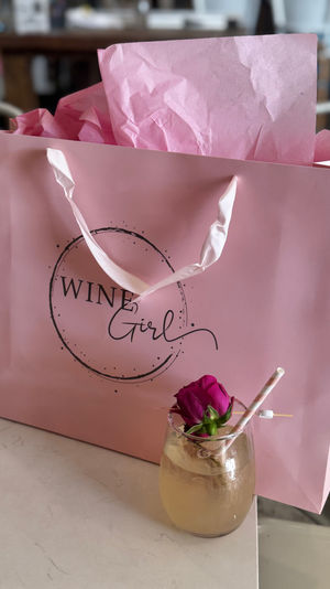 Pink gift bag with crinkled tissue paper and satin handles on a table, paired with a pale cocktail in a stemless glass garnished with a pink rose and striped straw — playful brunch gift scene