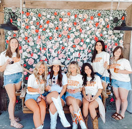 Seven women in matching graphic tees, denim shorts and cowboy boots posing with drinks on a wooden swing in front of a dense pink-and-white rose wall with neon script — sunny, fun girls' day photo