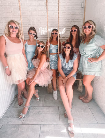 Seven friends in pastel dresses and heart-shaped sunglasses posing on indoor rope swings against a white brick wall with a neon sign — playful brunch or bachelorette photo spot.