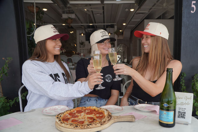 Three friends in baseball caps clinking champagne glasses over a wood-fired pepperoni pizza at a sunny outdoor café patio.