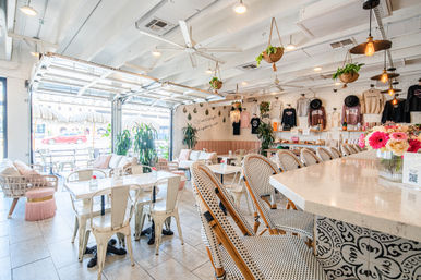 Bright, airy sunlit café interior with a marble bar and woven bistro chairs, hanging potted plants and Edison pendant lights, open garage-door seating, white brick wall and fresh floral centerpiece.