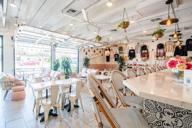 Bright, airy sunlit café interior with a marble bar and woven bistro chairs, hanging potted plants and Edison pendant lights, open garage-door seating, white brick wall and fresh floral centerpiece.