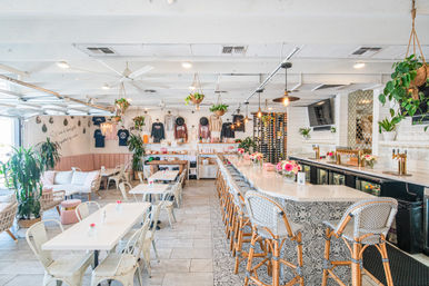 Sunlit modern café and wine bar interior with a long marble counter, woven barstools, hanging plants, wine rack and cozy white dining tables for a bright brunch atmosphere.