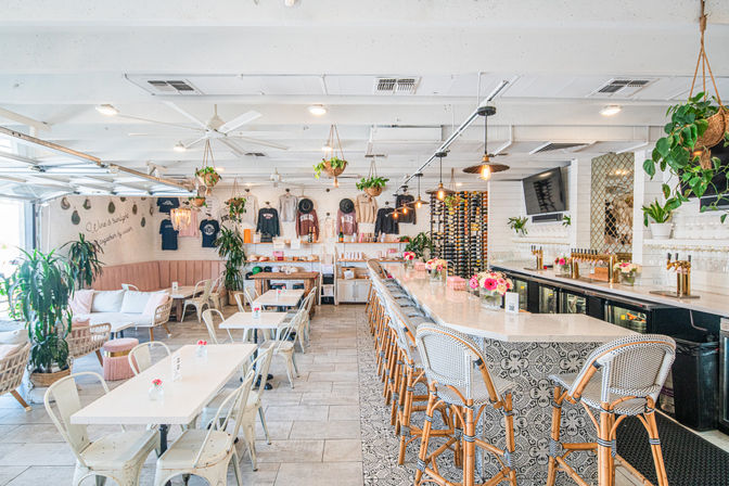 Sunlit modern café and wine bar interior with a long marble counter, woven barstools, hanging plants, wine rack and cozy white dining tables for a bright brunch atmosphere.