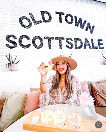 Smiling woman in a wide-brim hat toasting a glass over a wooden tasting flight in a pastel café with a white brick wall reading “Old Town Scottsdale.”