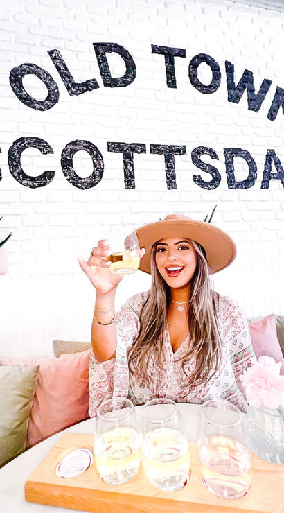 Smiling woman in a wide-brim hat raising a glass over a wooden tasting flight inside a bright cafe with “Old Town Scottsdale” painted on a white brick wall, pink pillows and succulents visible.