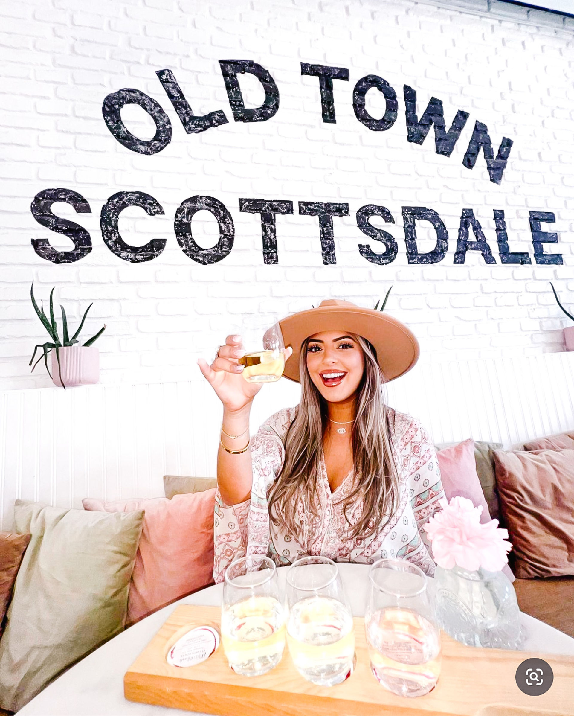 Smiling woman in a wide-brim hat raising a glass over a wooden tasting flight inside a bright cafe with “Old Town Scottsdale” painted on a white brick wall, pink pillows and succulents visible.