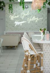 Chic Napa Valley wine bar interior with white floral wall and neon script sign, white-woven rattan bar stools at a marble counter, hanging greenery and soft pink floral accents.