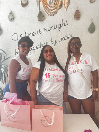 Three women in sunglasses posing indoors at a celebration, wearing white graphic T‑shirts (center shirt reads “46 Reasons for the Party!!”) with pink gift bags on a table and beaded wall decor and script lettering behind them.