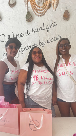 Three women in sunglasses posing indoors at a celebration, wearing white graphic T‑shirts (center shirt reads “46 Reasons for the Party!!”) with pink gift bags on a table and beaded wall decor and script lettering behind them.