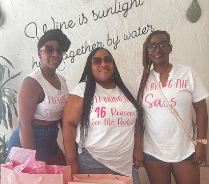 Three women in sunglasses posing indoors at a celebration, wearing white graphic T‑shirts (center shirt reads “46 Reasons for the Party!!”) with pink gift bags on a table and beaded wall decor and script lettering behind them.