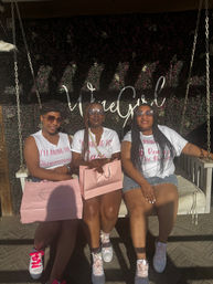 Three friends on a white swing at an outdoor patio with a green floral wall backdrop and cursive sign, wearing matching white tees and sunglasses, holding pink gift bags and pink-accented sneakers in a sunlit afternoon celebration.