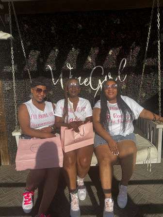 Three friends on a white swing at an outdoor patio with a green floral wall backdrop and cursive sign, wearing matching white tees and sunglasses, holding pink gift bags and pink-accented sneakers in a sunlit afternoon celebration.