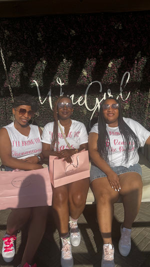 Three friends on a white swing at an outdoor patio with a green floral wall backdrop and cursive sign, wearing matching white tees and sunglasses, holding pink gift bags and pink-accented sneakers in a sunlit afternoon celebration.