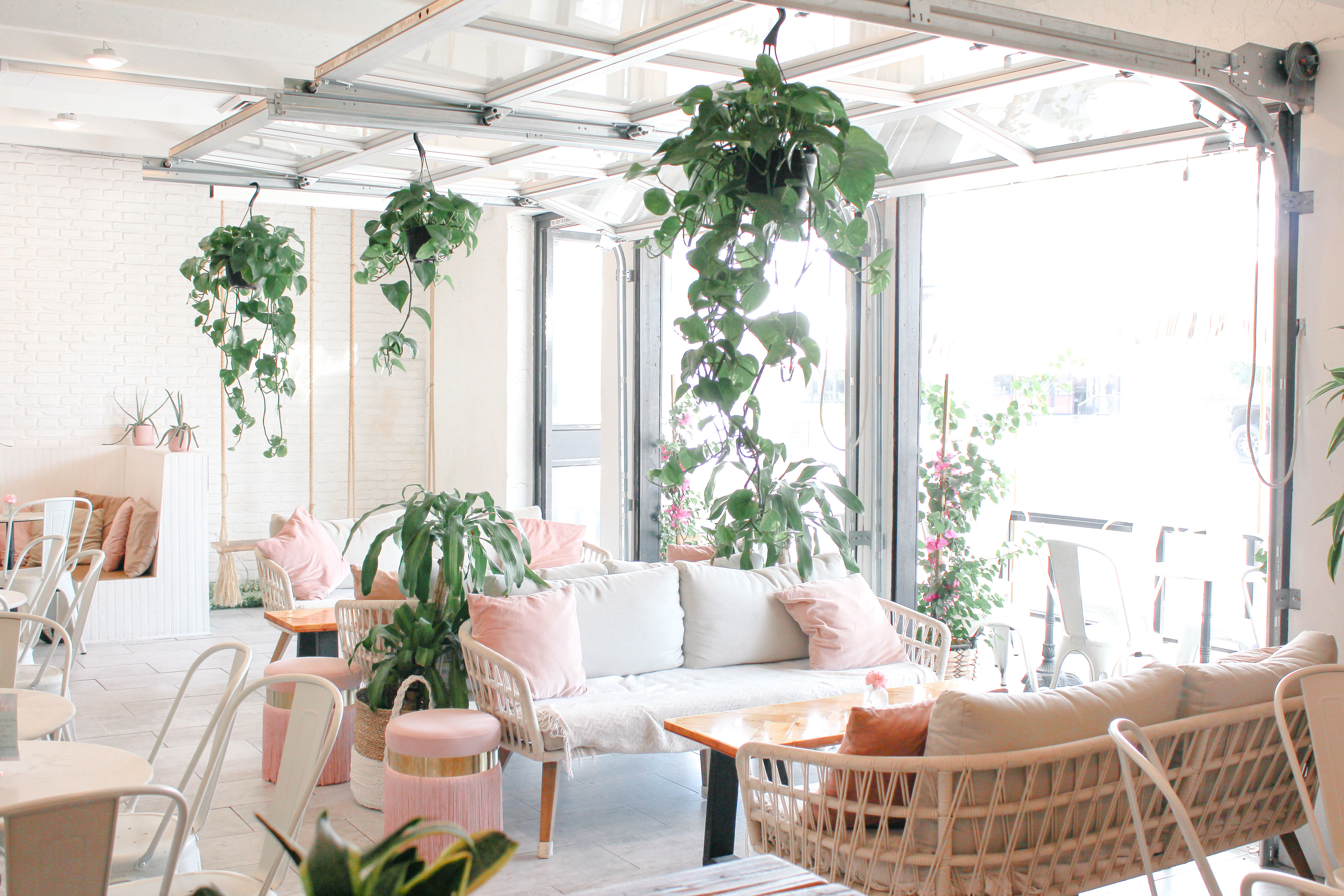 Sunlit modern cafe interior with open garage-style doors, rattan sofas, pastel pink cushions, wooden tables and cascading hanging plants creating a cozy, airy vibe.