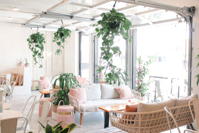 Sunlit modern cafe interior with open garage-style doors, rattan sofas, pastel pink cushions, wooden tables and cascading hanging plants creating a cozy, airy vibe.