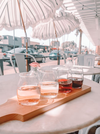 Sunny coastal cafe patio with striped umbrellas and palm-lined street in the background; a wooden paddle holds a flight of four small glasses with light to dark beverages on a round marble table.