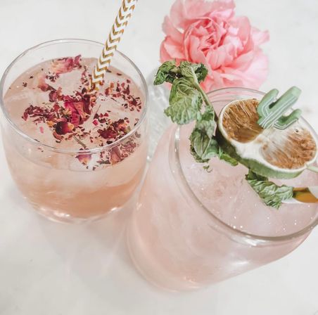 Blushing pink cocktails on a marble tabletop — one glass topped with dried rose petals and a gold chevron straw, the other with crushed ice, mint sprig and a dried lime wheel, pink peony in the background.