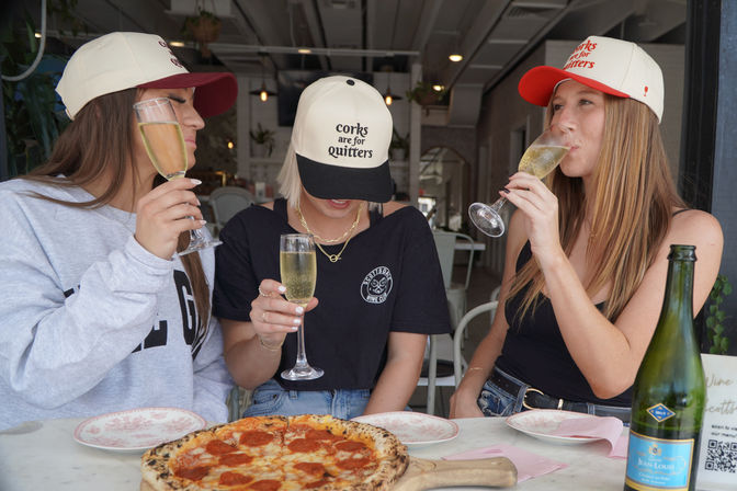 Three friends in baseball caps sipping champagne flutes over a pepperoni pizza at a cozy café patio — center cap reads "corks are for quitters", casual brunch and bubbly vibe.