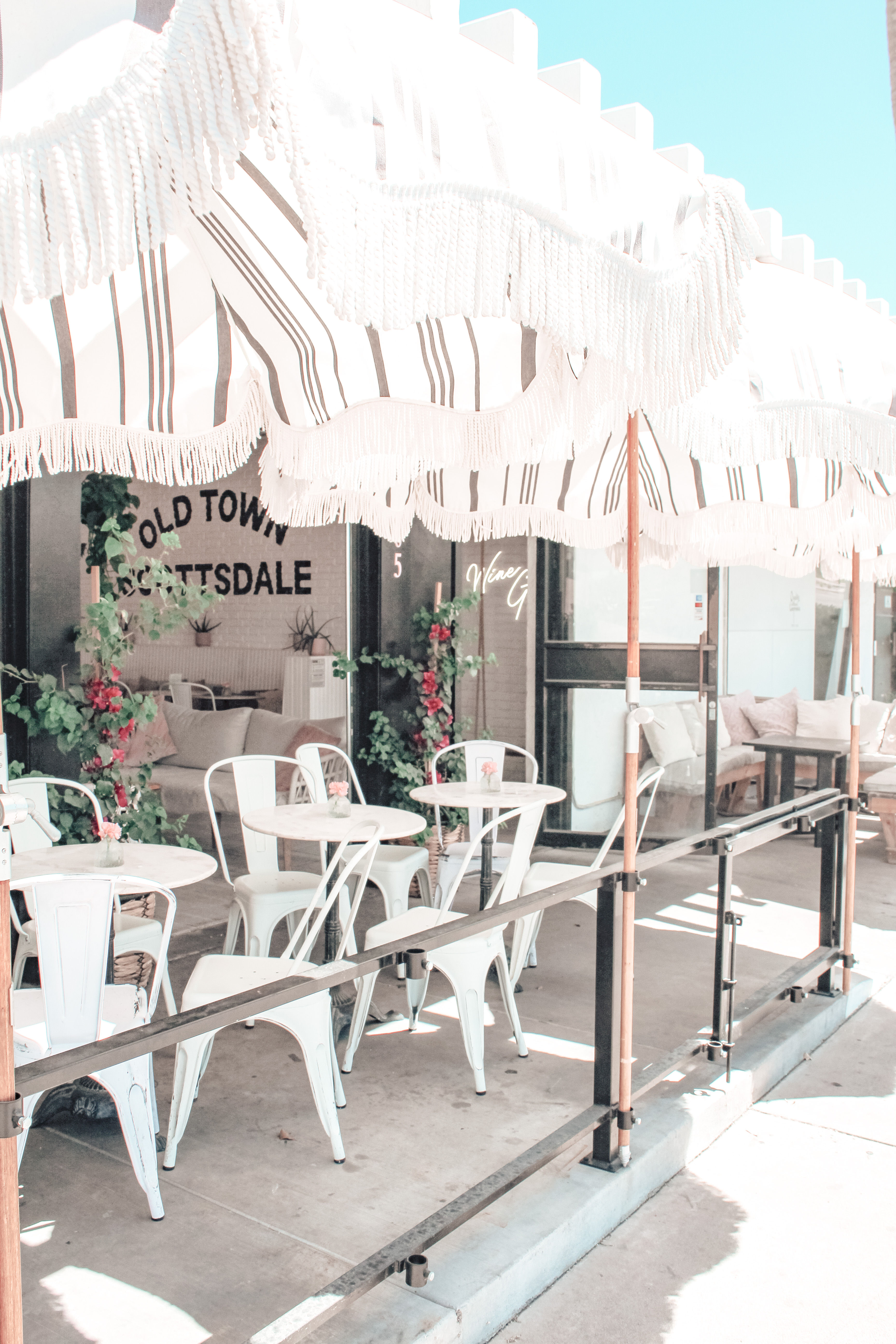 Sunlit outdoor café patio with striped fringed umbrellas, white metal bistro tables and chairs, small vases of flowers and potted greenery along a shaded sidewalk, pastel aesthetic.