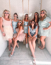 Seven friends in pastel dresses and rompers wearing pink heart-shaped sunglasses pose on rope swings against a white brick wall neon backdrop in a playful indoor photo-op