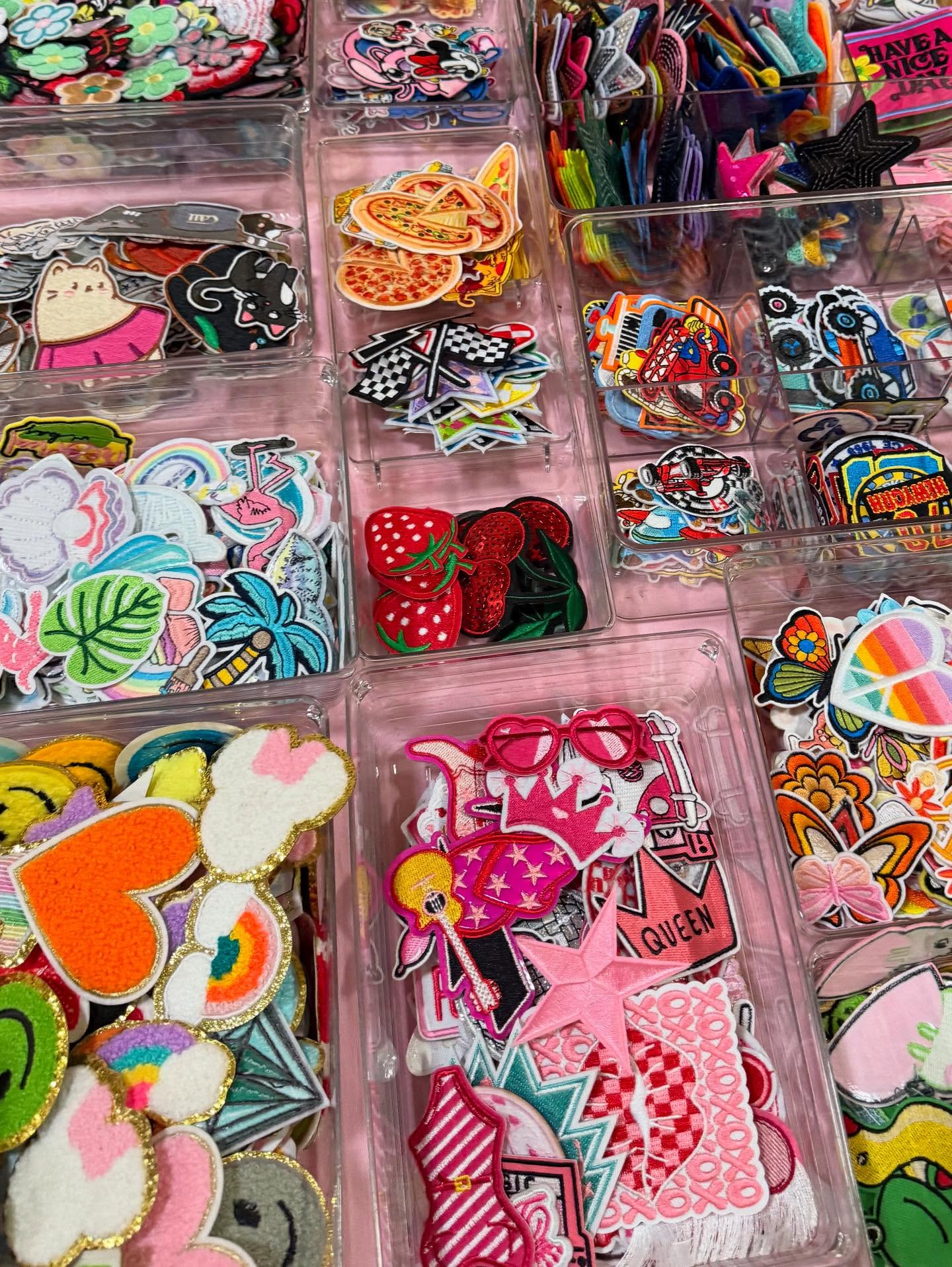 Vibrant assortment of embroidered iron-on patches—hearts, strawberries, pizza slices, palm leaves and stars—neatly sorted in clear plastic bins on a pink craft store display.