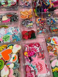Vibrant assortment of embroidered iron-on patches—hearts, strawberries, pizza slices, palm leaves and stars—neatly sorted in clear plastic bins on a pink craft store display.