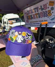 Bright purple trucker hat decorated with colorful Halloween patches (ghost, bats, spiderweb, candy, BOO) held in front of a sunny outdoor craft fair vendor tent and parked van.