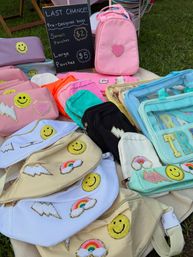 Colorful embroidered fanny packs, small backpacks and clear pouches with smiley faces, rainbows, hearts and lightning patches displayed on a blanket at an outdoor market