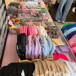 Bright accessory display of cute pastel zip pouches and coin purses on a pink table with clear bins of embroidered patches and stickers, plus stacked mesh headbands and colorful knit caps in a boutique-style setup.