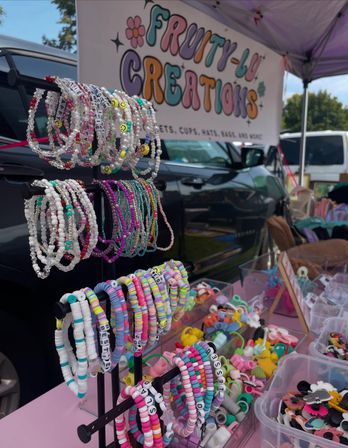 Colorful handmade beaded bracelets with pastel, neon, smiley and letter beads displayed on a rack and in bins at an outdoor craft market booth under a canopy.